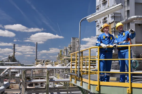 Blick von schräg oben auf Petrochemieanlage, rechts stehen diskutierend zwei Männer mit gelbem Helm und Blaumann auf einer Ballustrade
